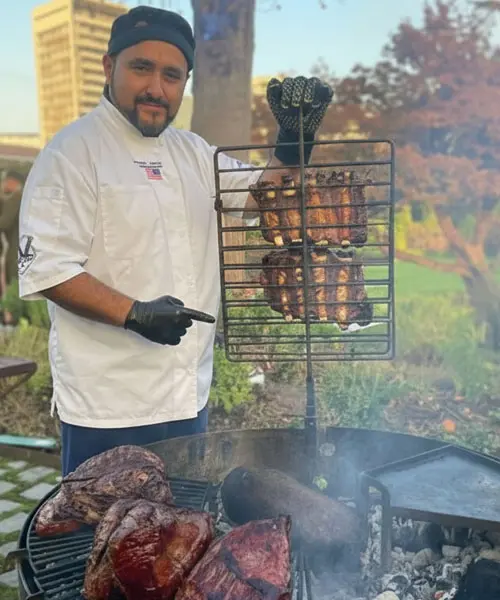 Jonathan Cárdenas realizando un asado argentino profesional en un evento, con carnes a la parrilla y asador a la cruz